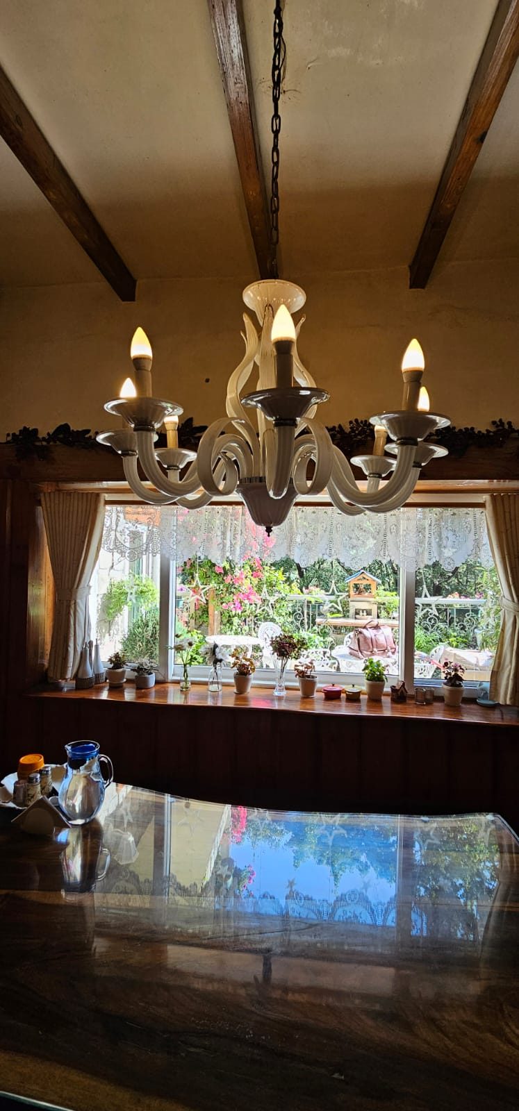 A cream chandelier above a polished wooden table, with a flower-lined window beyond