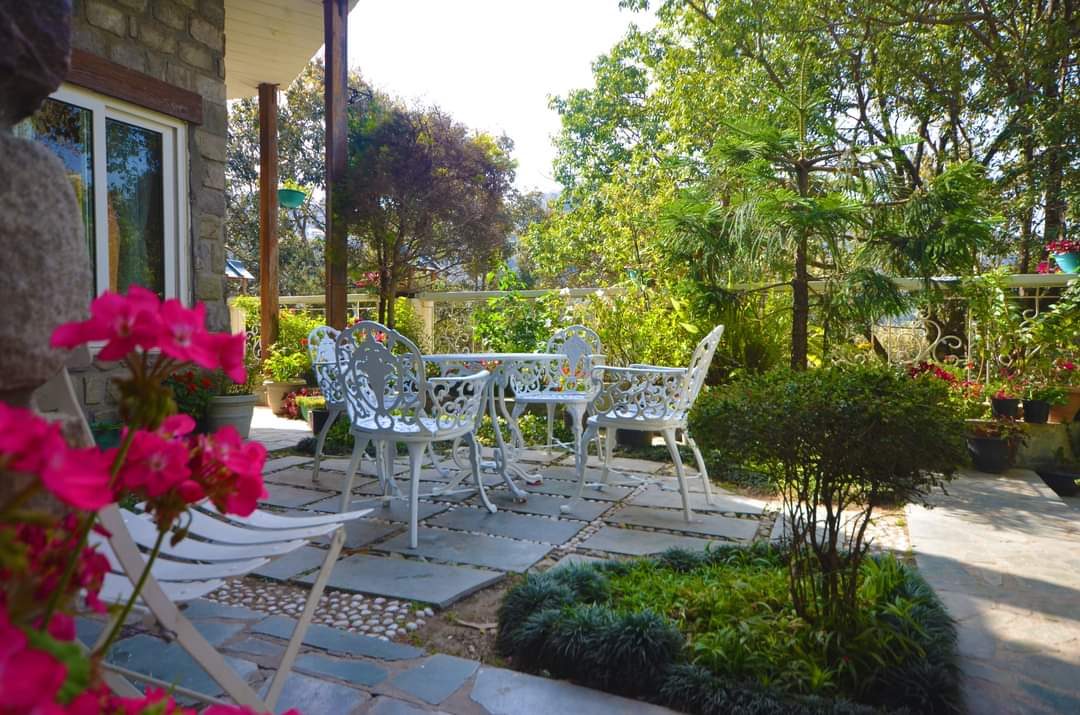 Stone-paved garden terrace with white wrought-iron chairs, pink geraniums and tall trees