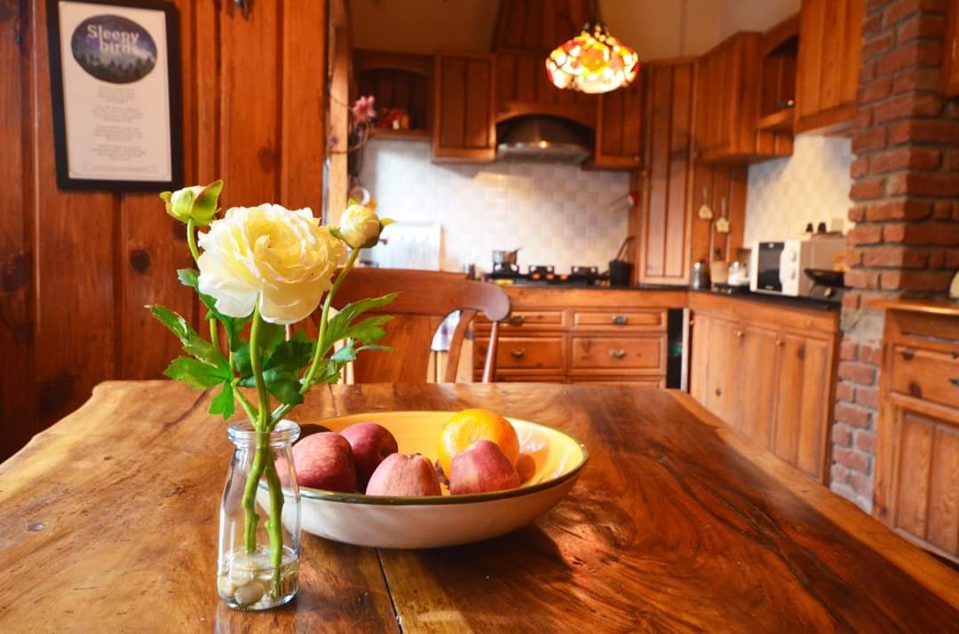 A white rose in a glass bottle and a bowl of fruit on a live-edge wooden table, with the kitchen behind